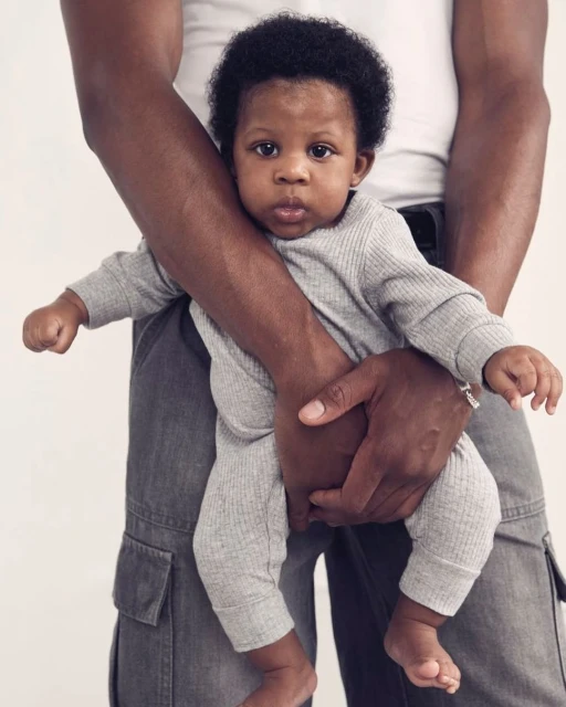 Infant in grey knit jumpsuit held by adult in front of white background. Casual, indoor studio shot emphasizing baby clothing.