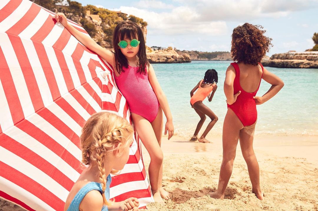 Four children in swimsuits at the beach with a striped beach umbrella, showcasing swimwear fashion and summer vacation.