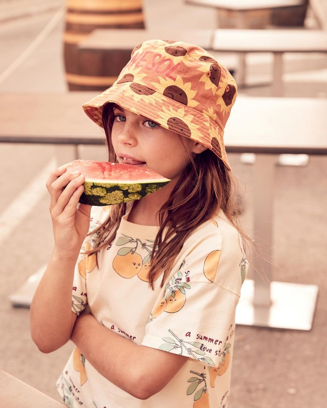 Young girl with a sunflower hat eats watermelon outside. She is wearing a patterned shirt, enjoying a refreshing snack, with a barrel visible.