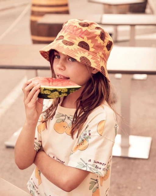 Young girl with a sunflower hat eats watermelon outside. She is wearing a patterned shirt, enjoying a refreshing snack, with a barrel visible.