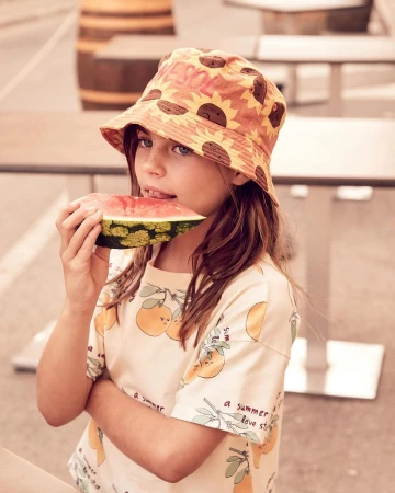 Young girl with a sunflower hat eats watermelon outside. She is wearing a patterned shirt, enjoying a refreshing snack, with a barrel visible.