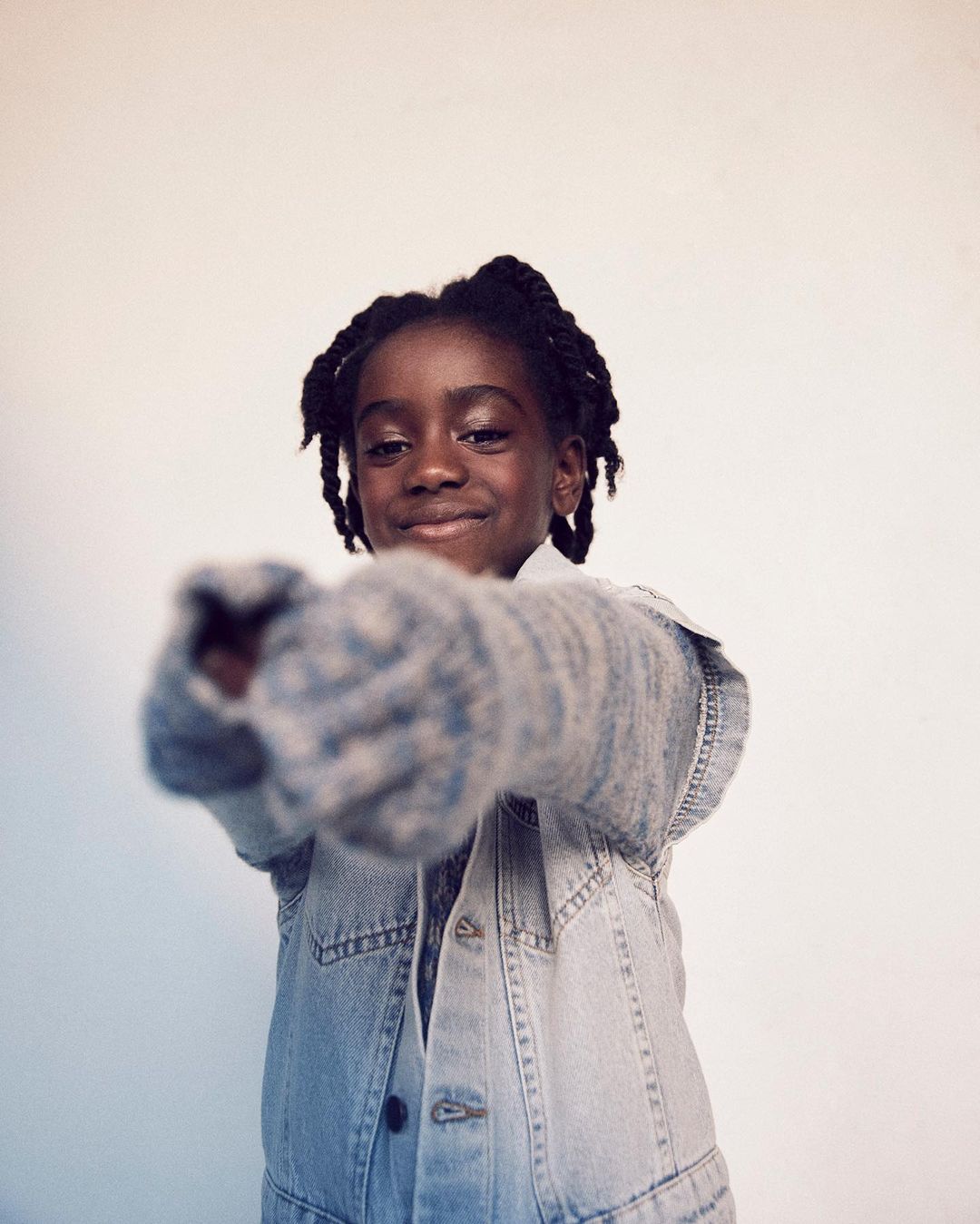 Smiling child with braids points towards camera. Child wears a light denim vest and gray knit sleeves against a light neutral backdrop.