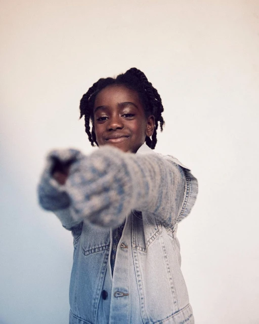 Smiling child with braids points towards camera. Child wears a light denim vest and gray knit sleeves against a light neutral backdrop.