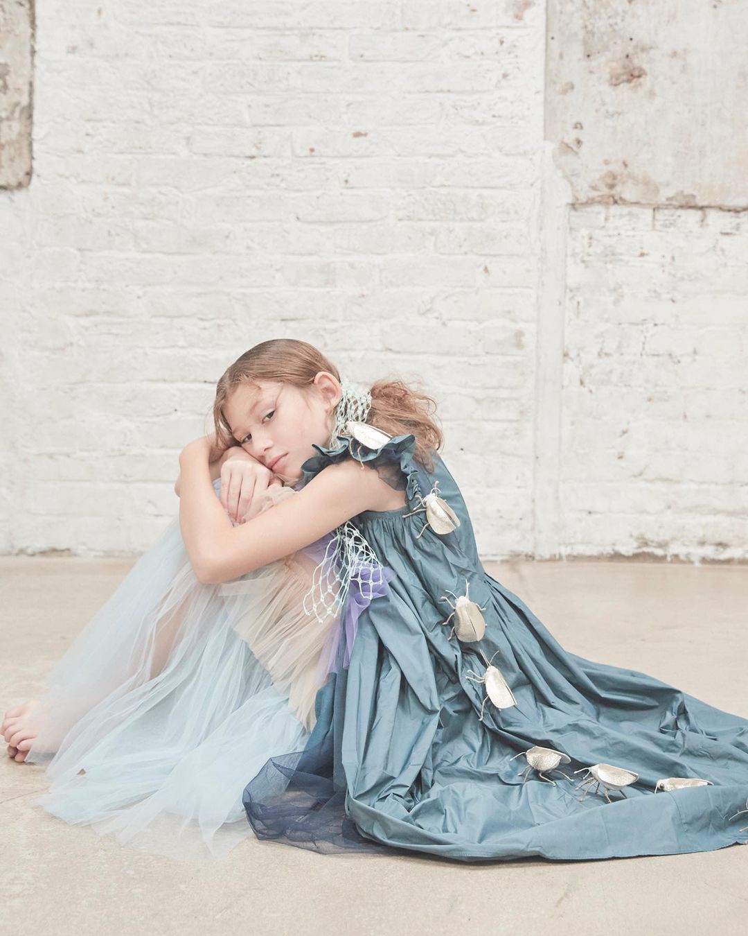 Young girl in a flowing blue dress with decorative beetle accents, seated against a textured white brick wall indoors. Dreamy aesthetic.