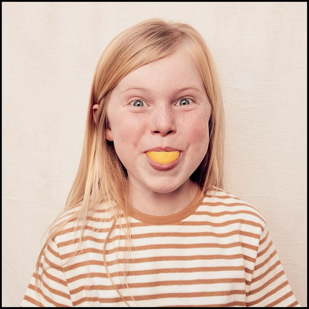 Close-up of a smiling blonde girl with freckles, wearing a striped shirt, holding a slice of lemon in her mouth against a plain background.