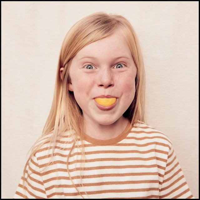 Close-up of a smiling blonde girl with freckles, wearing a striped shirt, holding a slice of lemon in her mouth against a plain background.