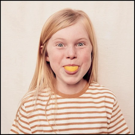 Close-up of a smiling blonde girl with freckles, wearing a striped shirt, holding a slice of lemon in her mouth against a plain background.