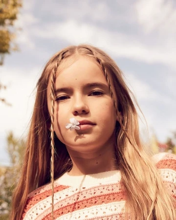 Young girl with braided long blonde hair holding a small white flower between her lips against a pale sky.