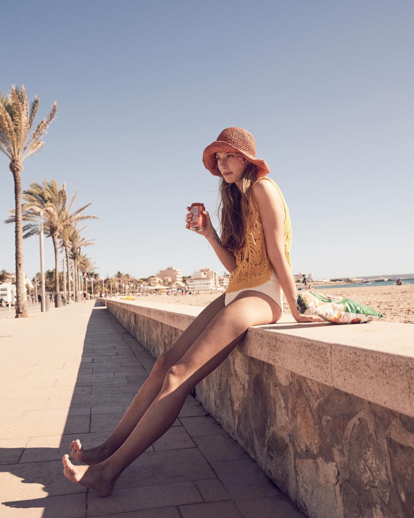 A young woman sits on a wall at the beach holding a drink, wearing a yellow knit top and hat, with palm trees in the background.