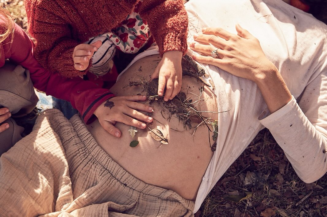 Pregnant woman lying outdoors with two children playing with leaves and twigs on her stomach, in a natural light setting.