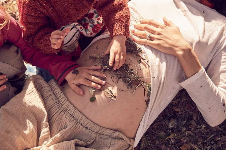 Pregnant woman lying outdoors with two children playing with leaves and twigs on her stomach, in a natural light setting.