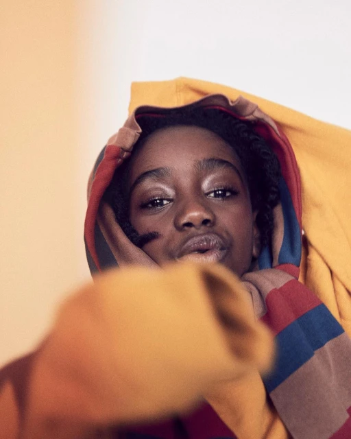 Close-up studio portrait of a young Black woman with hand near her face, wearing a hooded, striped shirt against a peach-colored background.
