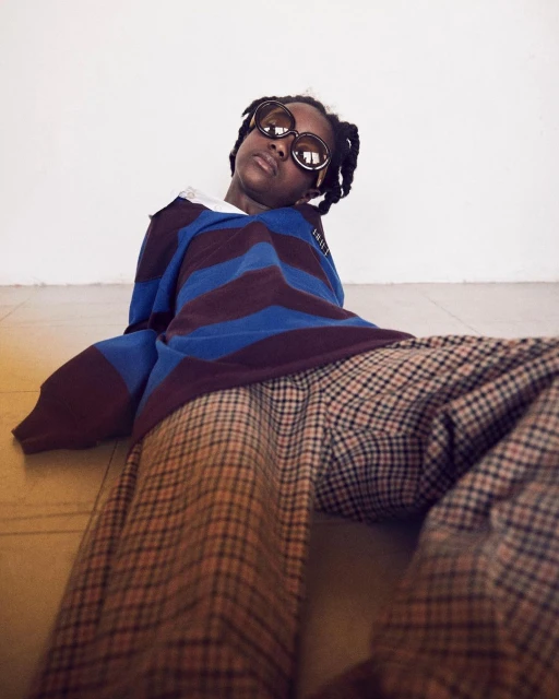 Person with round sunglasses sits on floor wearing a striped rugby shirt and plaid pants against a stark white wall in a portrait.