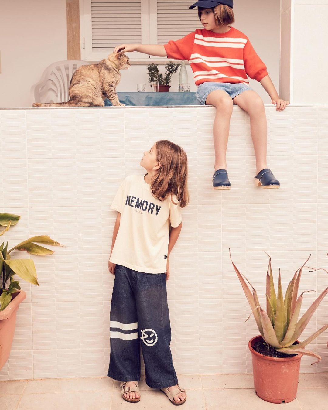 Two children, one petting a cat, sit on a tiled ledge, and the other stands below wearing a t-shirt and wide leg pants, in an outdoor setting.