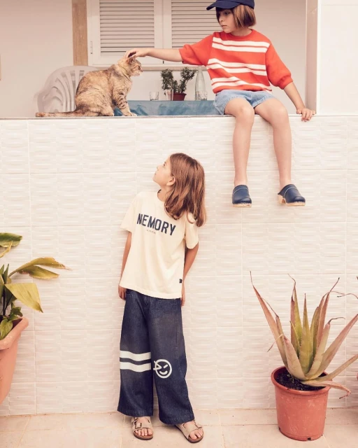 Two children, one petting a cat, sit on a tiled ledge, and the other stands below wearing a t-shirt and wide leg pants, in an outdoor setting.