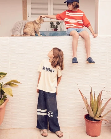 Two children, one petting a cat, sit on a tiled ledge, and the other stands below wearing a t-shirt and wide leg pants, in an outdoor setting.