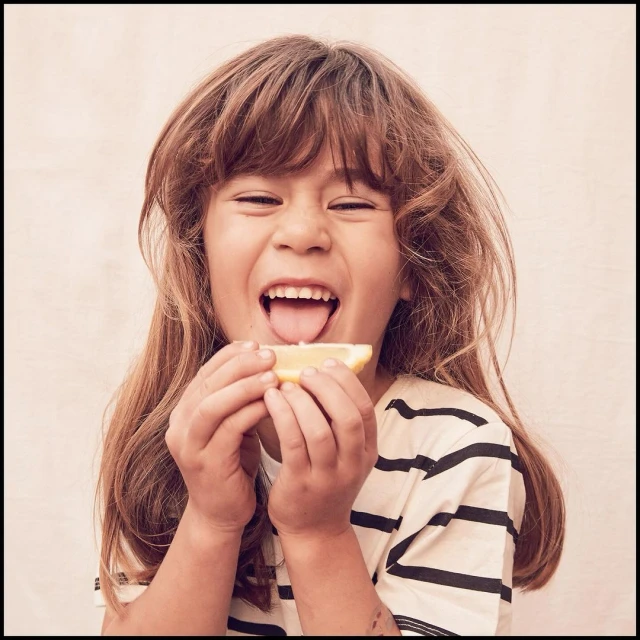 Child with long brown hair tasting a slice of lemon, with an open mouth and eyes closed, wearing a striped shirt against a beige background.
