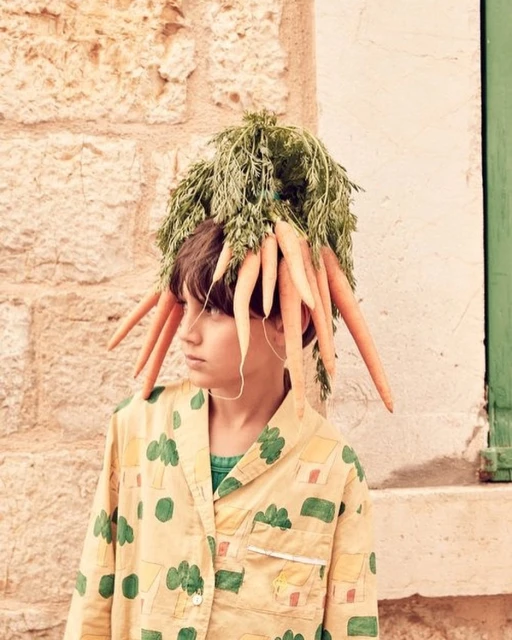 A young boy poses outdoors wearing a playful hat made of fresh carrots and greenery, adorned in a patterned pyjama top.