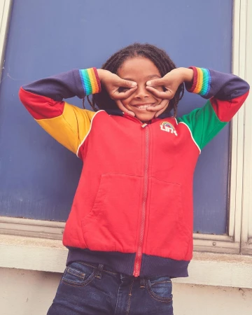 Young child wearing a colour-blocked zip-up hoodie making circles with hands over eyes against a blue backdrop.
