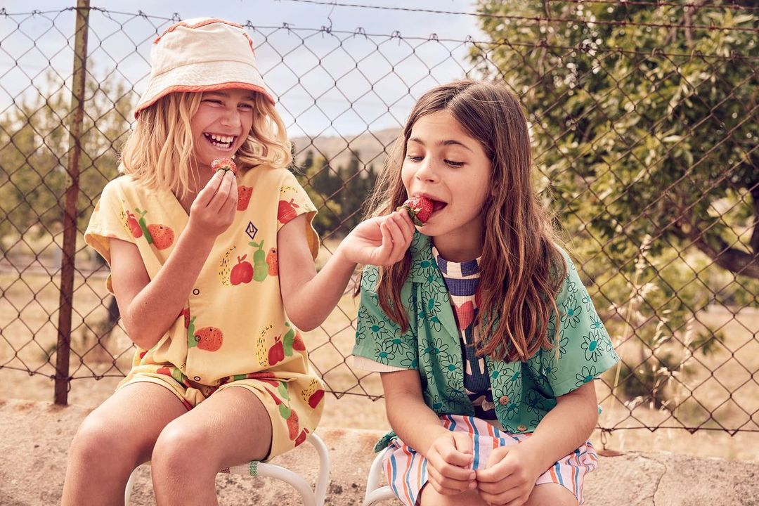 Two young girls, one with blonde hair and a bucket hat, the other with brown hair, sit side by side and eat strawberries outdoors.
