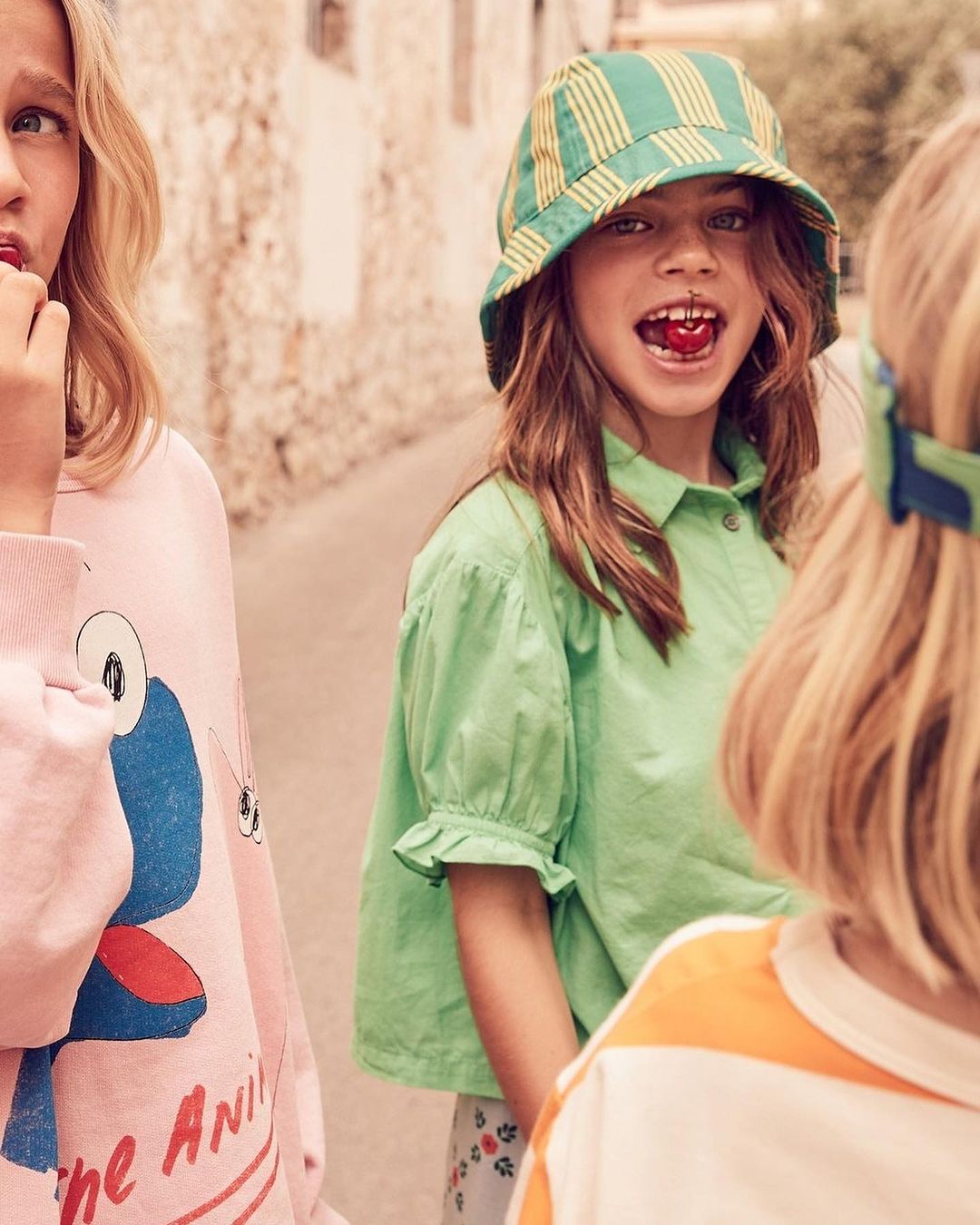 Three children in casual clothing are standing outdoors, one in a striped hat eating a cherry, in a medium shot, during daytime.