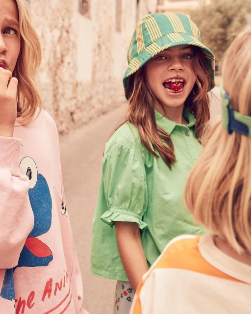 Three children in casual clothing are standing outdoors, one in a striped hat eating a cherry, in a medium shot, during daytime.