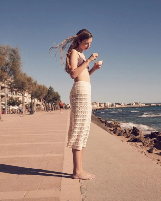 Young woman in swimsuit top and crochet skirt standing barefoot eating ice cream on a boardwalk by the ocean under a clear blue sky.