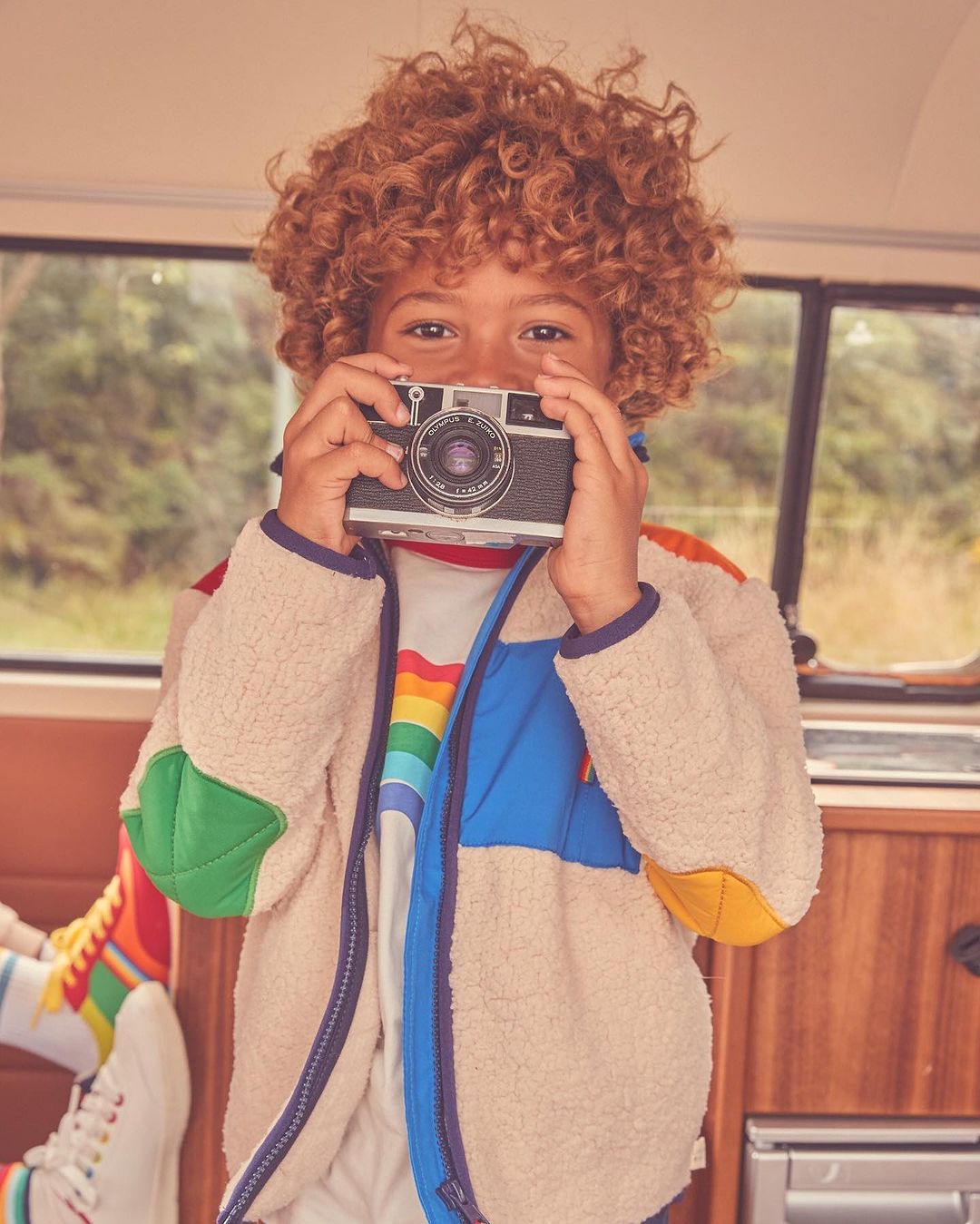 A child with curly red hair holds a vintage camera, smiling indoors, wearing a colorful jacket and rainbow shirt, inside a vehicle.