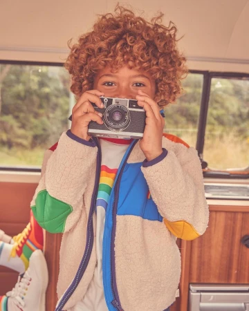 A child with curly red hair holds a vintage camera, smiling indoors, wearing a colorful jacket and rainbow shirt, inside a vehicle.