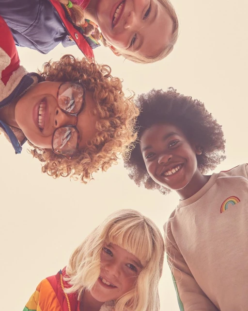 Four smiling children huddle closely together, looking down at the camera with bright expressions in an eye-level, close-up composition.
