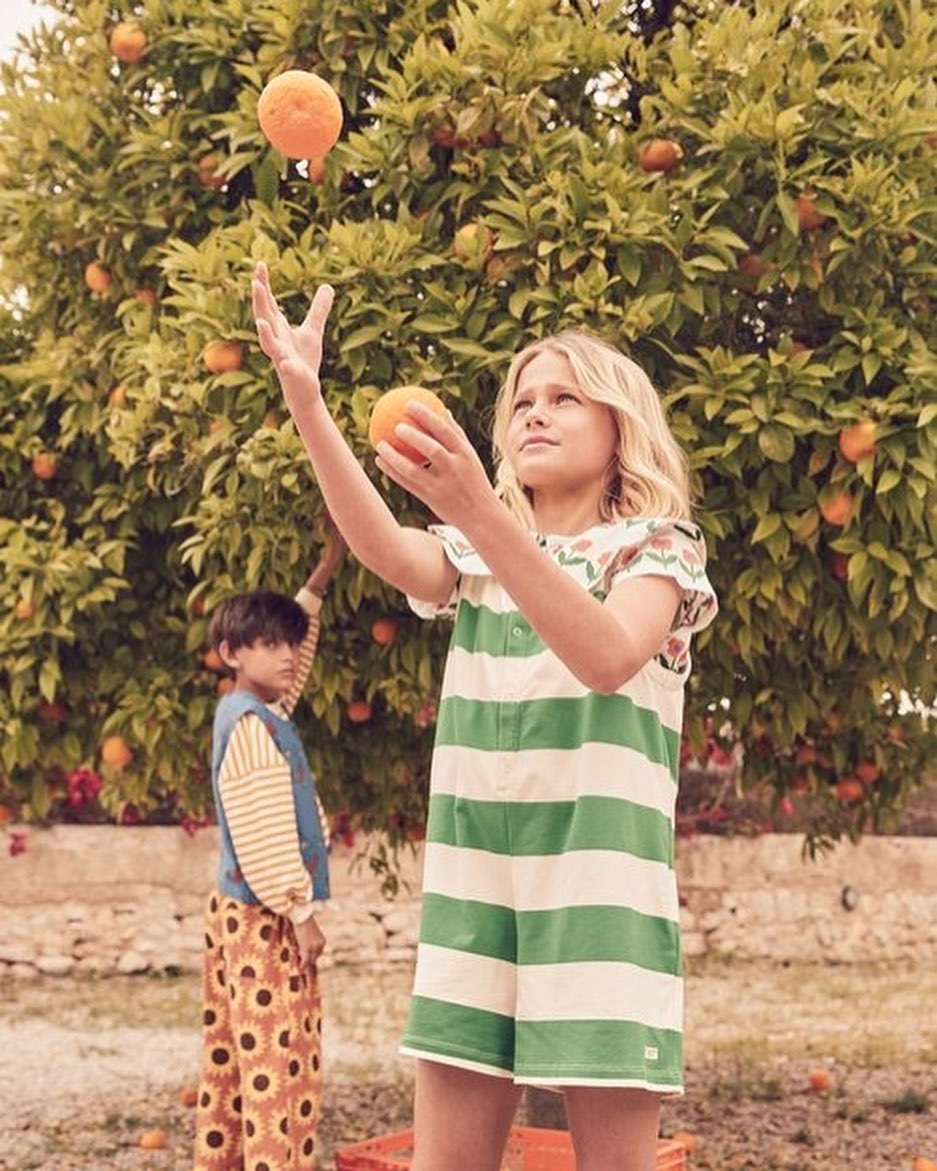 A young girl tosses an orange in the air, standing outdoors near a laden orange tree, with another child visible in the background.