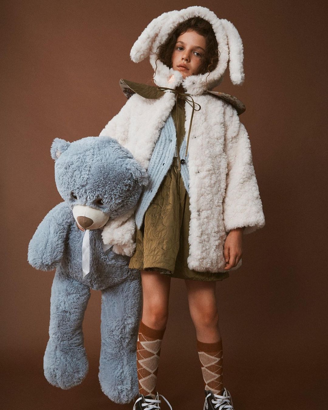 Young child with a blue plush teddy bear in a studio shot. The child is wearing a hooded bunny ear coat and brown argyle socks.