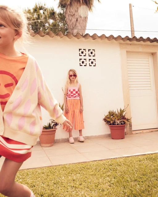 Two young girls are playing in the backyard near a white wall with potted plants in the summer sunshine, dressed in casual clothing.