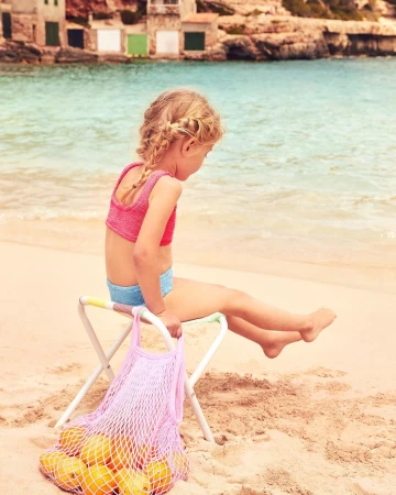 Young girl with braided hair sitting on a stool at the beach with a net bag of oranges, near blue water and coastal buildings.