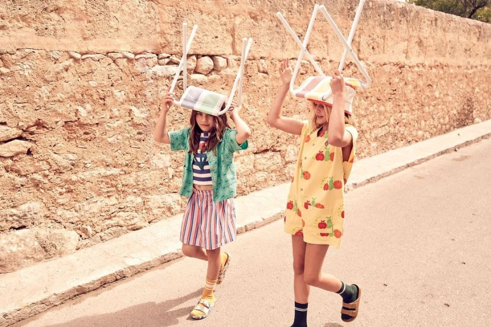 Two young girls in colourful summer outfits playfully balance chairs on their heads while walking on a sunny outdoor street.
