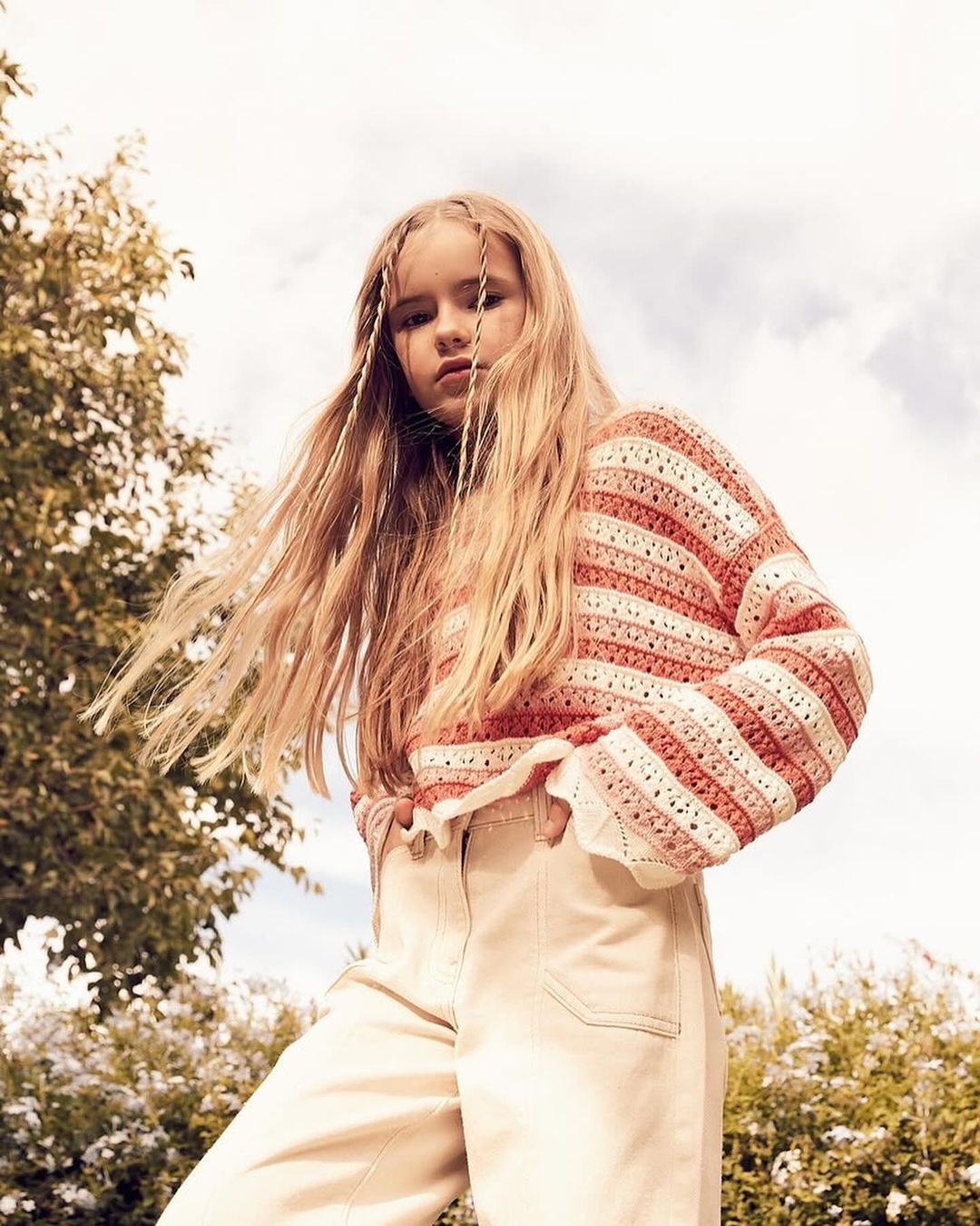 Child model with long blond hair poses in a light pink and white striped knit sweater and light beige pants outdoors against sky.