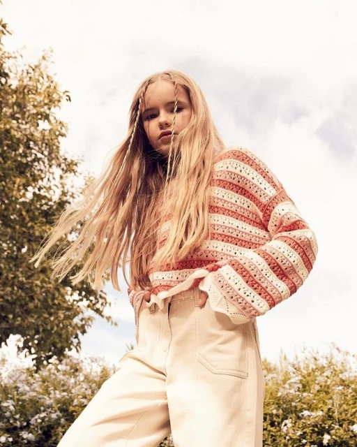 Child model with long blond hair poses in a light pink and white striped knit sweater and light beige pants outdoors against sky.