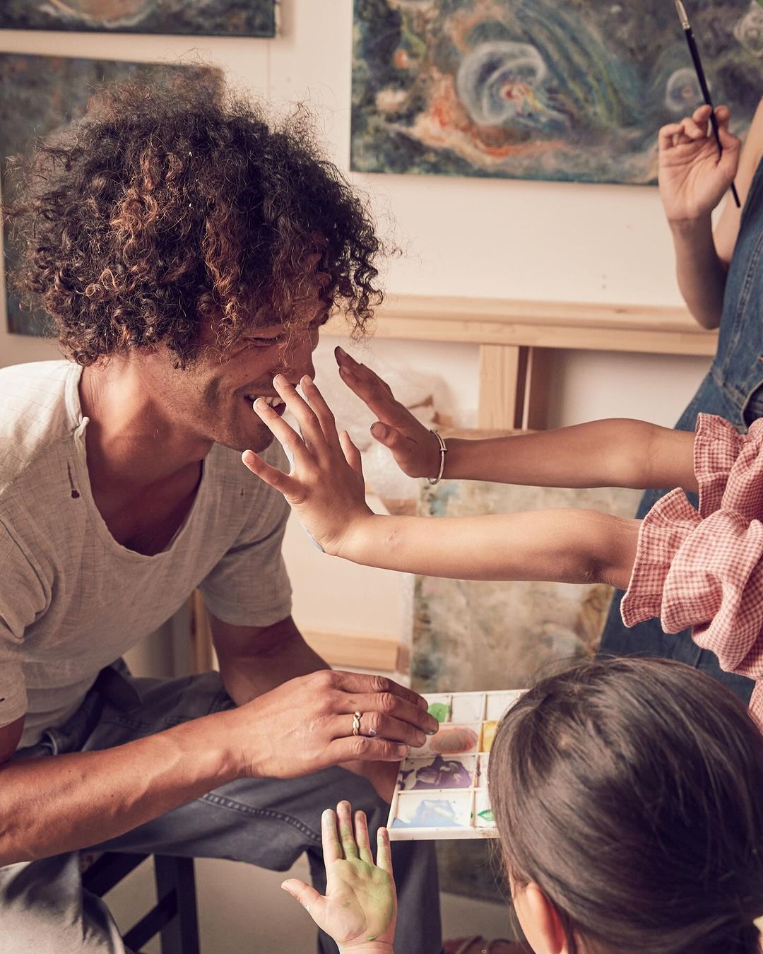 Family painting together in a brightly lit art studio with paintbrushes, watercolor paints, and art canvases visible.