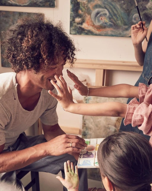 Family painting together in a brightly lit art studio with paintbrushes, watercolor paints, and art canvases visible.