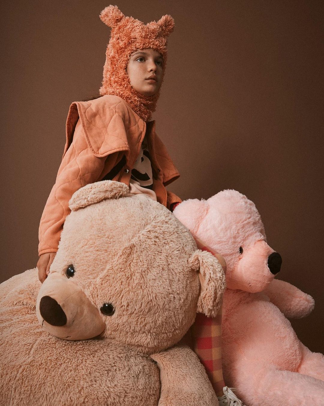 Young person wearing a coral fleece hat and quilted jacket sits on an oversized light brown teddy bear with a pink teddy bear behind.