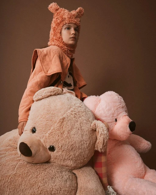 Young person wearing a coral fleece hat and quilted jacket sits on an oversized light brown teddy bear with a pink teddy bear behind.