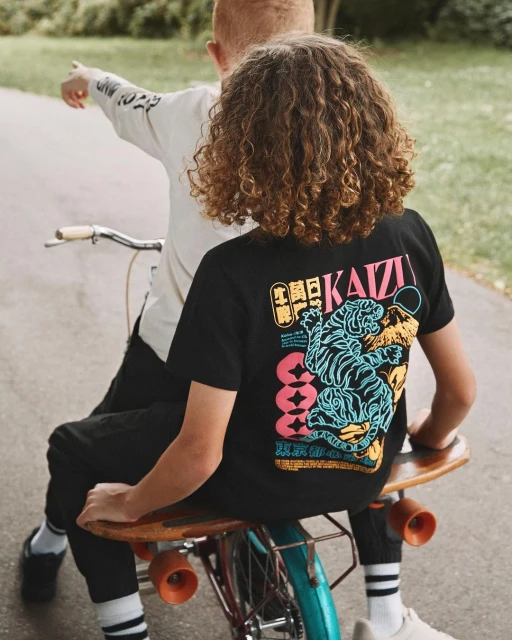 Two children ride together on a bicycle. One wears a black t-shirt with a tiger design, the other a cream-colored t-shirt pointing ahead.