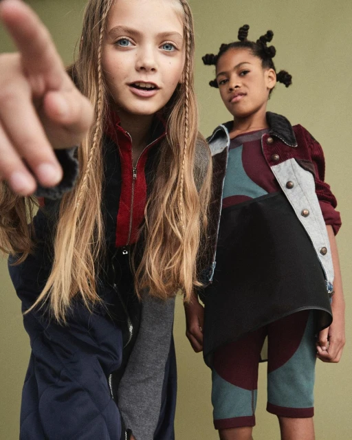 Two young girls pose in stylish patterned jackets in an eye-level shot. The girl in front points to the camera with a neutral facial expression.
