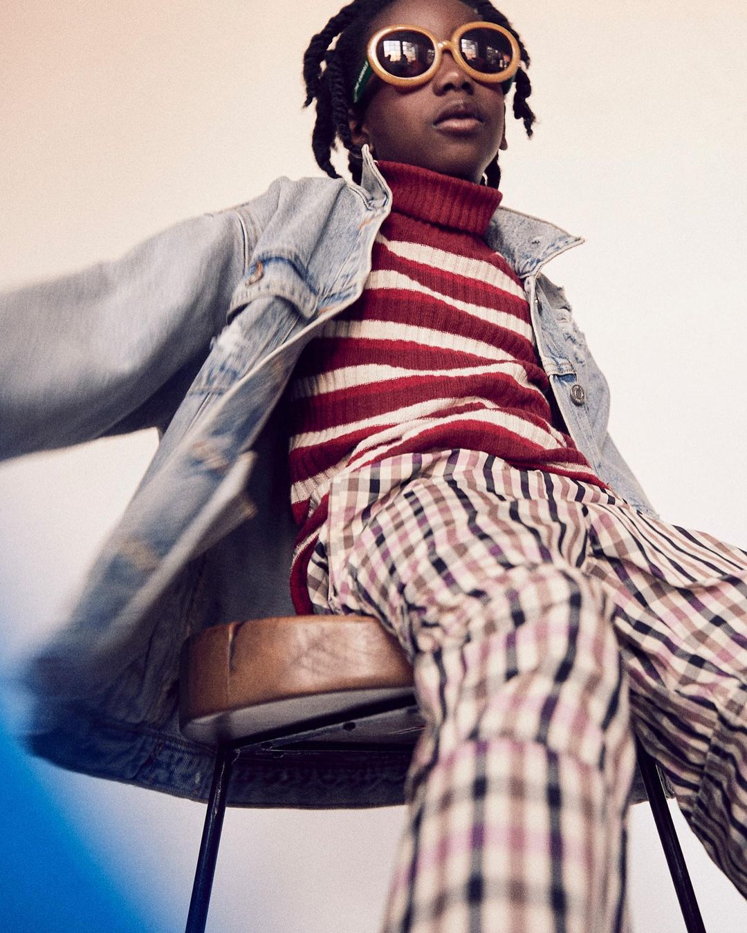 A young person with stylish eyewear and denim jacket, seated on a stool, gazes confidently in a studio environment, highlighting youth fashion.