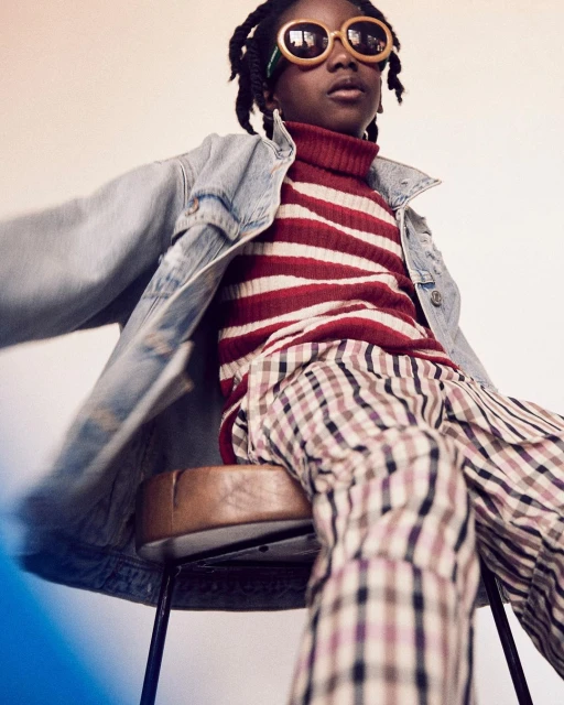 A young person with stylish eyewear and denim jacket, seated on a stool, gazes confidently in a studio environment, highlighting youth fashion.