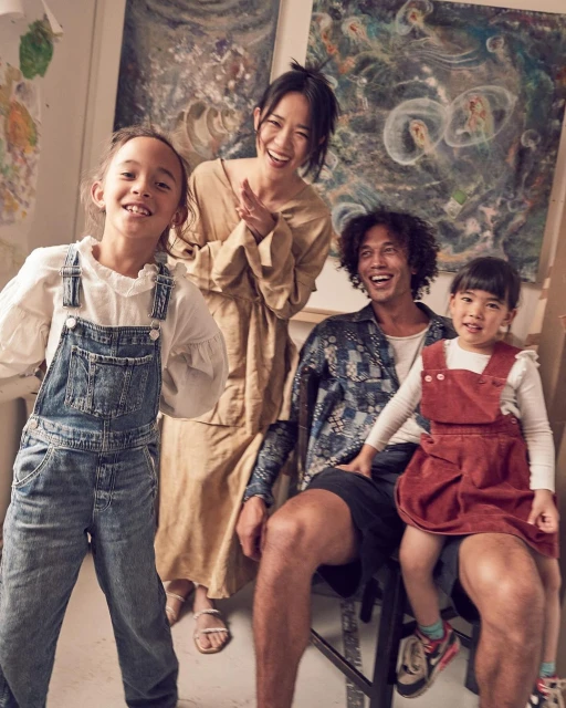 A happy family of four poses together in an art studio with smiles, featuring two young girls, their mother in a dress, and their father.