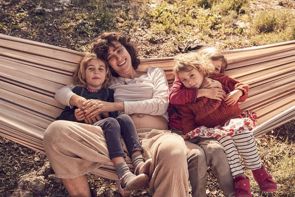 A mother with her three young children sitting in a hammock outdoors. The children are cuddling with their mother in a daytime shot.