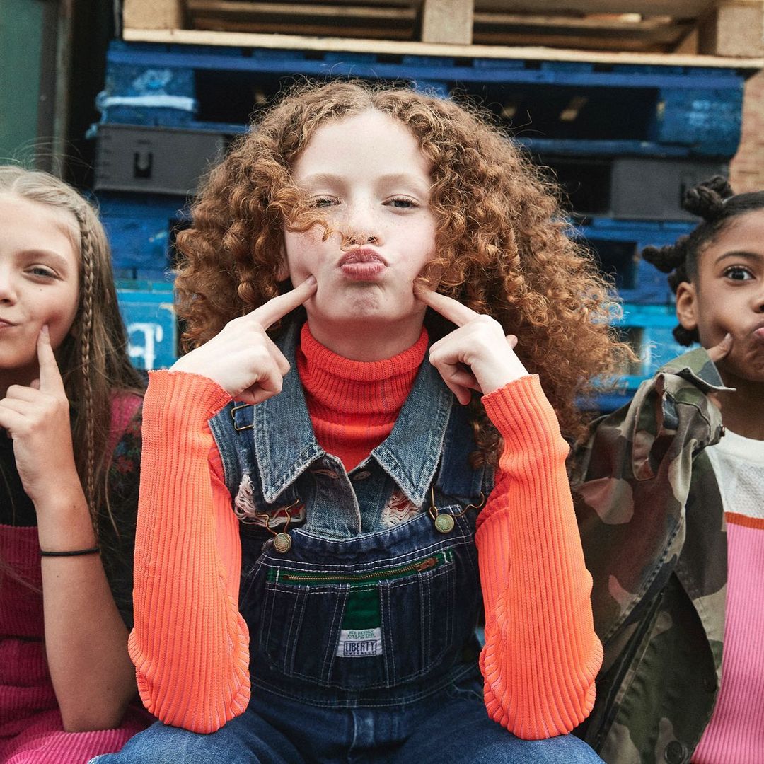 Three girls pose cheek-to-cheek in front of a pallet wall, one with red hair in denim overalls and an orange turtleneck sweater.