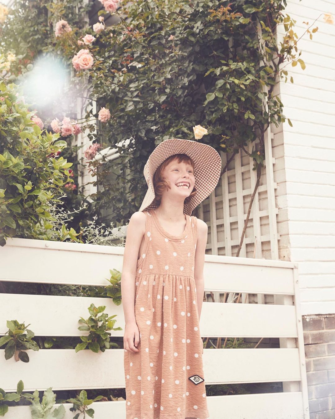 A smiling young girl in a polka-dot dress and sunhat stands in front of a white fence with pink roses climbing on a white-sided house.