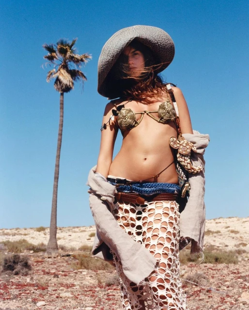 A woman poses in a bikini top and skirt near a palm tree against a blue sky in a desert landscape, exuding a sun-kissed, carefree vibe.